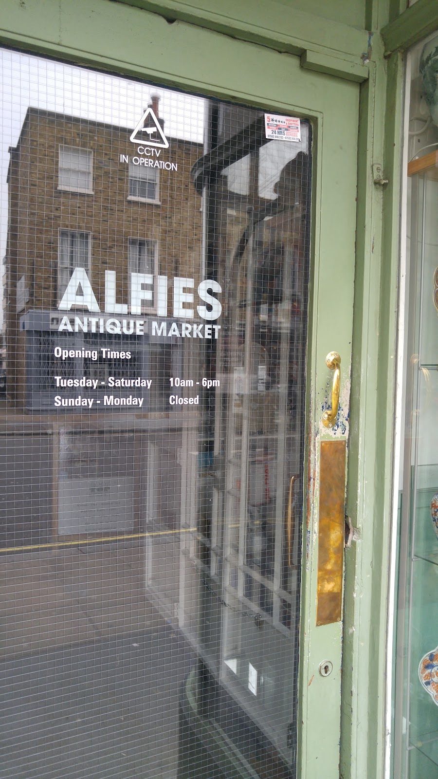 The green-framed glass entrance door of Alfies Antique Market on Church Street, with the brick street reflected in the glass